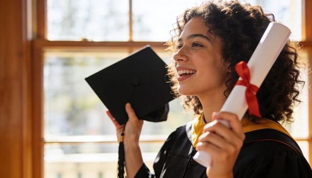 Smiling graduate holding diploma and cap celebrating academic achievement at sunlit window