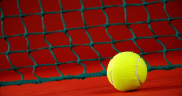 Close-up view of a tennis ball resting beside a vibrant red court with green netting in the background. Ideal for use in sports, competition, training themes, tennis advertisements, articles, and promotional materials.