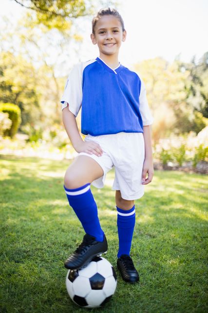 Young girl wearing soccer uniform standing in park with one foot on soccer ball. Ideal for use in sports-related content, youth activities promotions, fitness and health campaigns, and outdoor recreation advertisements.