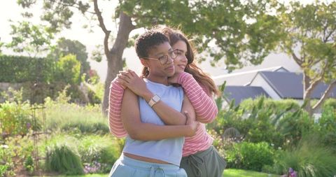 Two diverse women embracing in sunlit backyard garden, casual clothes and smartwatch