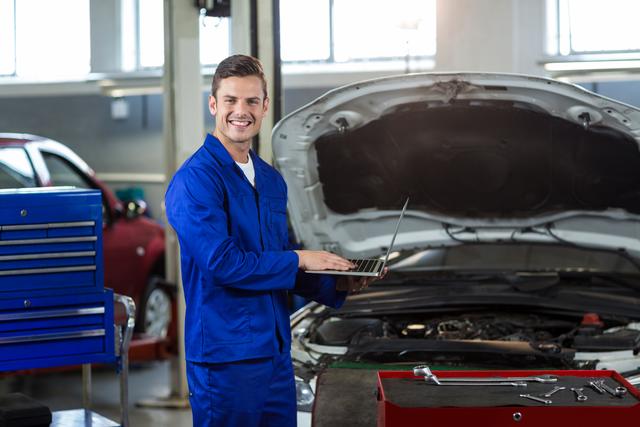 Mechanic wearing a blue jumpsuit smiling while using a laptop in an auto repair garage. Could be used for articles on auto repair, technology in automotive industry, or promotional material for car service centers.