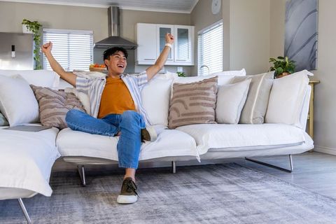 Young asian man relaxing and celebrating on white sectional sofa in modern open-plan living room
