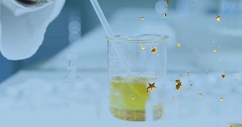Gloved hand using pipette over graduated beaker with yellow liquid and gold star confetti