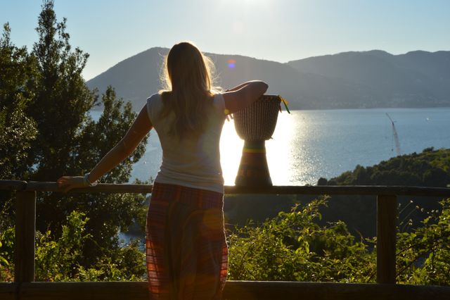 Woman standing by wooden fence, enjoying serene lake view and mountains in background during sunset. Silhouette emphasizes peaceful and reflective moment. Ideal for travel blog, meditation and relaxation themes, promoting outdoor activities, or emphasizing serenity and contemplation.