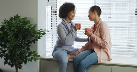 Diverse couple sharing a cozy moment by window with hot beverage