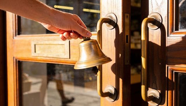 Ringing brass service bell at sunlit wooden shop doors with brass handles and reflections