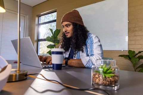 Typing remote worker at home desk with travel mug, plants and whiteboard in daylight