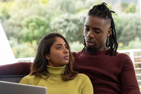 Intimate multiracial couple relaxing on sofa with laptop near sunlit window