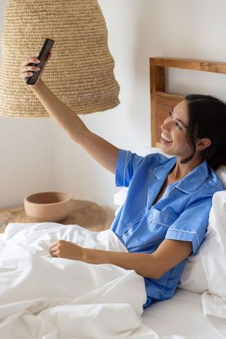 Smiling woman taking selfie in blue pajamas on cozy bed under woven lamp