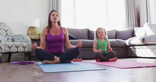 Mother and daughter practicing meditation on yoga mats in a cozy living room. Both engaged in a mindfulness exercise, seated cross-legged with eyes closed. Ideal for content related to family wellness, home workouts, mental health, and bonding activities.