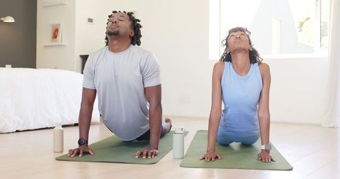African american couple practicing morning yoga on mats in sunlit bedroom with wearables