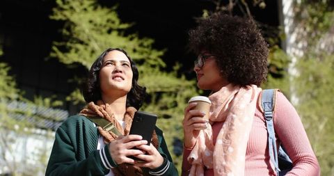 Two women walking in sunlit urban park holding smartphone and coffee, chatting casually
