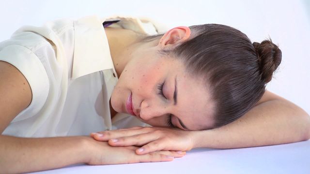 Young woman sleeping on a table with her hands resting under her head, against a white background. Depicts rest, relaxation, and taking a break. Useful for wellness, mental health, relaxation products, and restful living concepts.