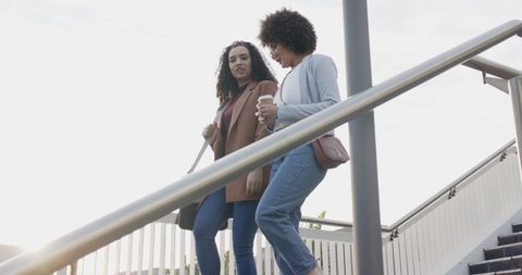 Two professional women walking down sunlit urban stairs talking while carrying coffee