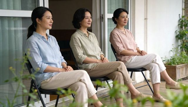 Three asian midlife women sitting on porch in pastel shirts relaxing on wooden deck