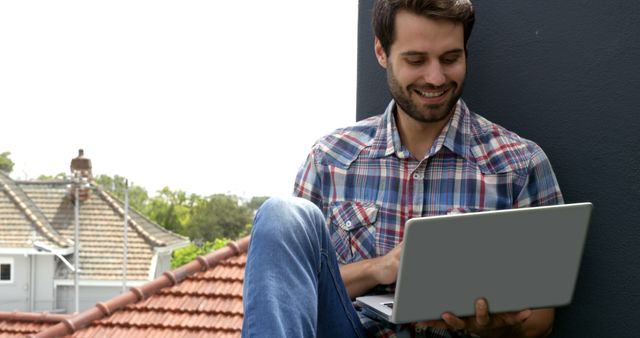 Young man casually using a laptop while sitting on a rooftop. Ideal for illustrating remote work, urban lifestyles, freelance jobs, relaxation in outdoor settings or tech use in non-traditional workspaces. This image can be used in promotions for remote work tools, lifestyle blogs, and social media content related to casual modern work environments.