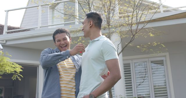 Portrait of happy diverse gay male couple with new house keys embracing in garden. Home, property, celebration, gay, relationship, togetherness, domestic life and lifestyle, unaltered.