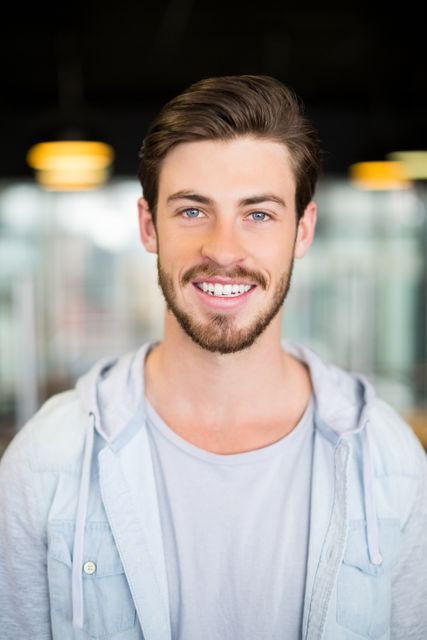 Young male executive smiling confidently in a modern office environment. Ideal for use in business-related content, corporate websites, professional networking profiles, and promotional materials showcasing a positive and approachable company culture.
