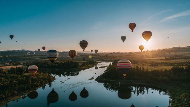 Colorful hot air balloons drifting over river valley at sunrise with reflections