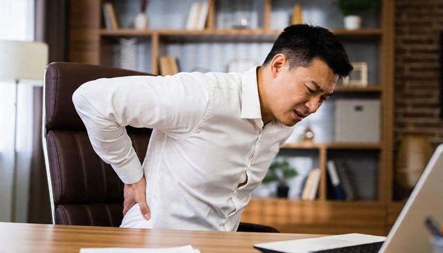 Middle-aged asian man holding lower back in office chair experiencing back pain from desk work
