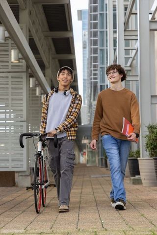 Two college friends walking with bicycle along modern urban campus corridor holding notebook