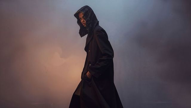 Moody high-fashion model wearing embellished hooded black trench coat, cinematic portrait