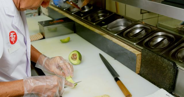 Chef in professional kitchen slicing avocado on cutting board with knife. Ideal for illustrating culinary skills, food preparation, and restaurant kitchen environments. Can be used for cooking websites, kitchen safety information, and culinary arts training materials.