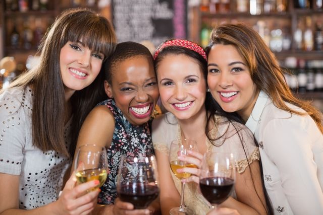 Group of diverse female friends smiling and holding wineglasses in a pub. Ideal for use in advertisements, social media posts, or articles about friendship, social gatherings, and leisure activities.