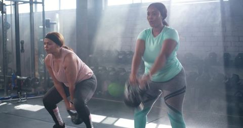 Diverse women swinging kettlebells in sunlit gym for strength training and partner workout