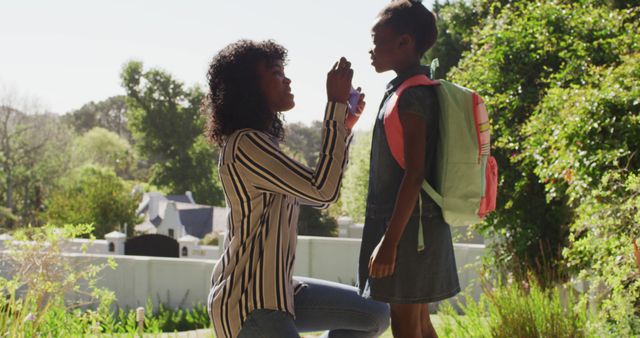 Mom kneeling and applying sunscreen to her daughter who is ready for school with a backpack. Useful for topics on family morning routines, childcare, healthy skincare habits, and parental care.