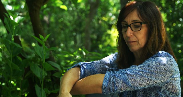 Mature woman sitting in a green forest, looking pensive. Her thoughtful expression reflects a moment of contemplation. Great for themes of solitude, relaxation, mindfulness, and nature therapy.