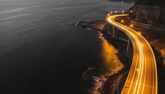 Curving coastal viaduct casting golden light trails along ocean cliffs at night