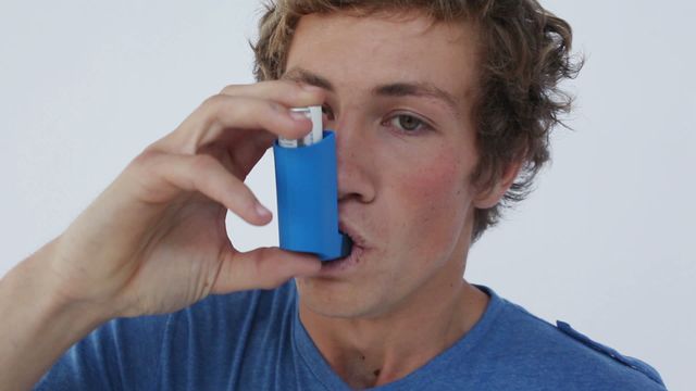 A man uses an inhaler to manage an asthma attack. Wearing a blue shirt, he is focusing on relieving his symptoms. This is ideal for promoting health education, illustrating chronic conditions management, or supporting healthcare-related articles.