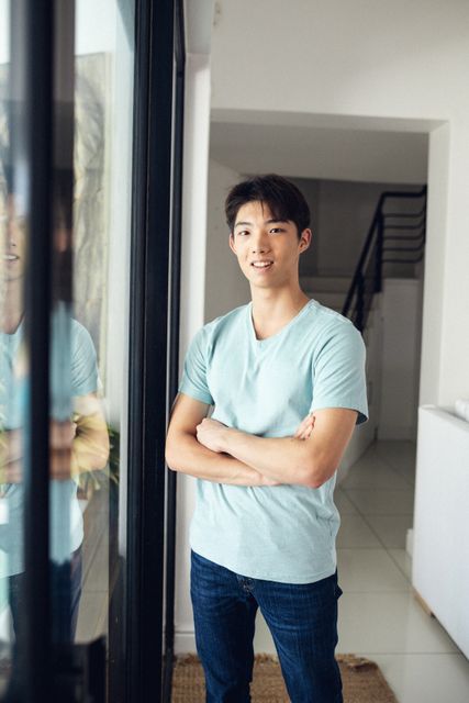 Portrait of smiling asian teenage boy with arms crossed standing by window at home, copy space. Unaltered, people, lifestyle and home concept.