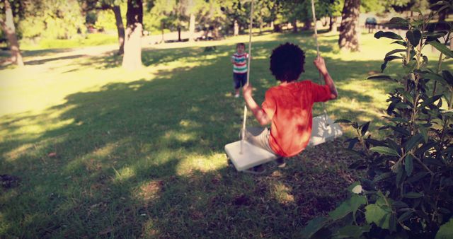 A young African American child enjoys a swing in a sunny park setting, with copy space. Another child watches from a distance, creating a sense of carefree childhood play.
