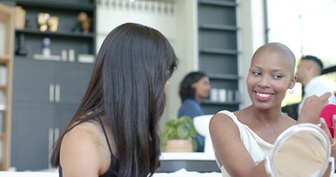 Smiling bald woman checking makeup with friend in contemporary studio apartment
