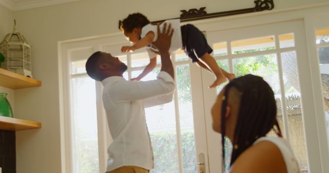 Father lifting young child in bright living room, with mother looking on. Suitable for themes of family, joy, indoors, parenting, or carefree living. Effective for articles, advertisements, social media posts showing family moments or positive home environments.
