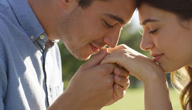 Romantic couple holding hands showing engagement ring leaning close in sunlit park