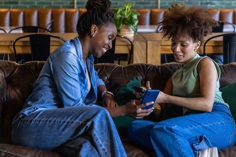 Diverse friends sharing smartphones and laughing on leather sofa in cozy cafe lounge wearing denim