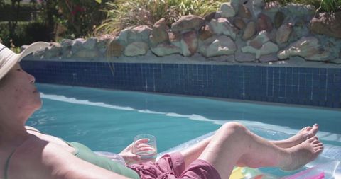 Senior asian woman reclining on inflatable pool float holding drink in sunny backyard