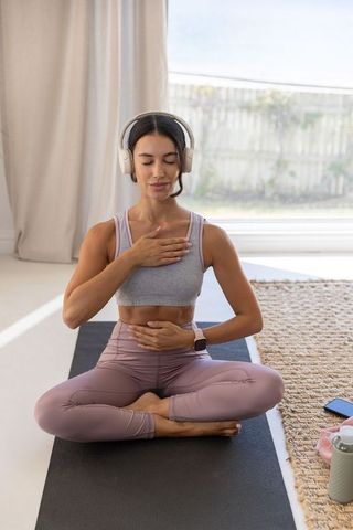 Young woman practicing mindful breathing at home with headphones and smartwatch on yoga mat