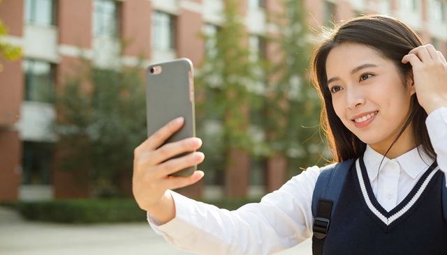 Smiling female student taking selfie on university campus with backpack and earbuds