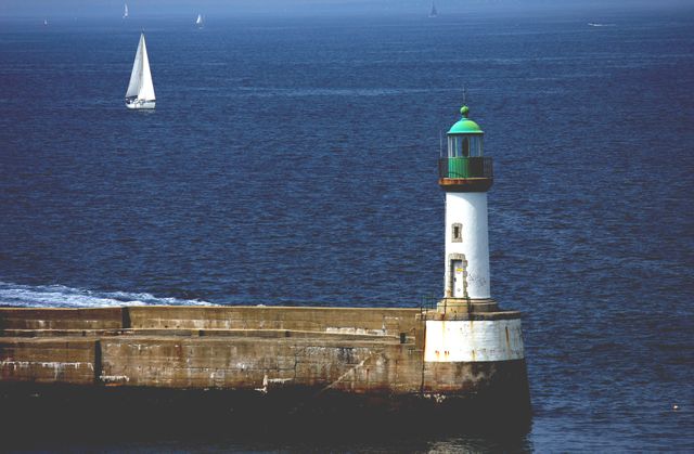 Lighthouse stands on a coastal pier with clear blue sea and sailing boats in the background. Useful for seaside travel promotions, sailing and maritime themes, coastal navigation guides, or as a calming desktop background.