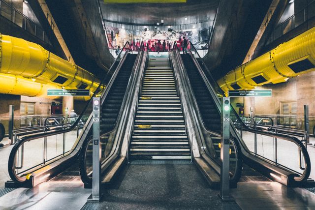 Central platform of a modern subway station featuring large yellow ducts and escalators leading up. Suitable for use in urban design, public transport infrastructure projects, and cityscape studies.