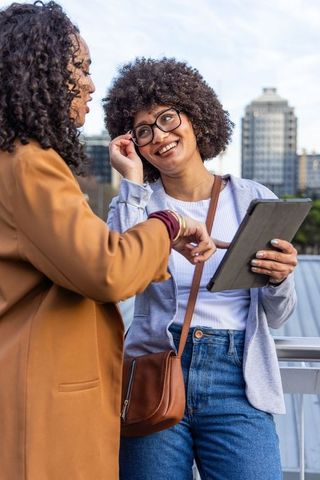 African american women chatting, sharing tablet and smiling on urban rooftop