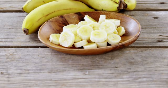 Sliced bananas are presented in a wooden bowl on a rustic table, with whole bananas in the background. Bananas, both whole and sliced, are often used in culinary preparations for their sweet flavor and nutritional benefits.