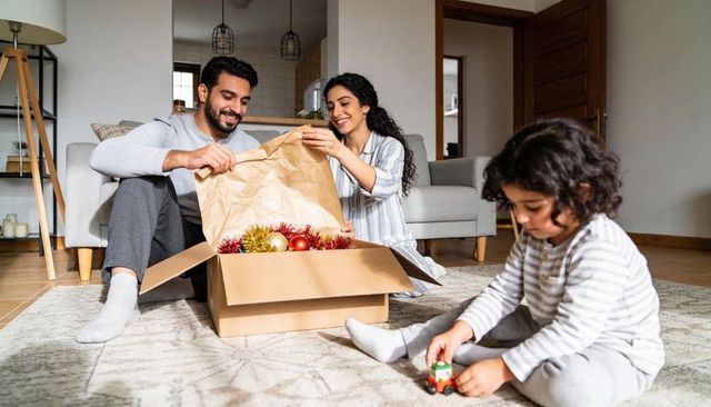 Family unpacking holiday ornaments on living room rug, parents smiling, child playing
