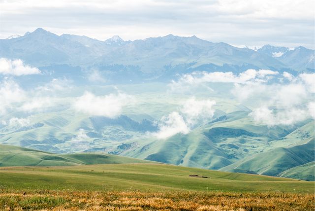 This detailed landscape with rolling green hills and mountains partly obscured by cloud coverage captures the beauty and tranquility of nature. Ideal for travel and nature-related content, this scene best represents remote mountain valleys and serene outdoor escapes. Perfect for advertising campaigns, websites focusing on outdoor activities, travel blogs, calendar images or desktop wallpapers.