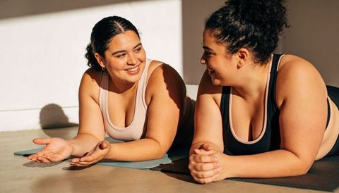 Plus-size women smiling and chatting on yoga mats in sunny indoor workout body positive