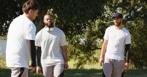 Multiracial friends standing and talking in park after workout wearing headphones and cap